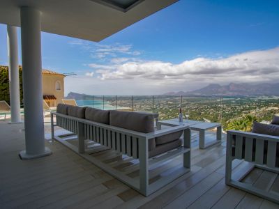 Expansive covered terrace with light decking and modern white lounge furniture, framing panoramic sea, coastal, and mountain views through a glass railing.