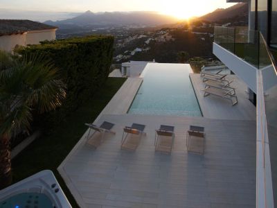 Expansive infinity pool with a light-toned deck, multiple sun loungers, and a separate jacuzzi, all set against a panoramic mountain sunset backdrop.