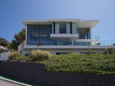 Exterior view of a modern three-level villa featuring extensive glass facades and glass railings, elevated above a stone retaining wall and lush vegetation by a curved road.