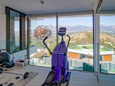 Gym with floor-to-ceiling windows showcasing panoramic mountain and valley views. Features an elliptical machine, free weights, and a glass balustrade in a modern setting.