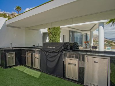 L-shaped outdoor kitchen featuring black granite countertops, dual sinks, a covered BBQ grill, and integrated stainless steel appliances, with reflected pool views.