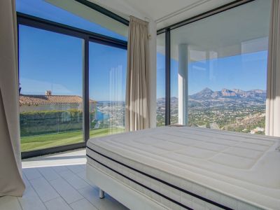 Modern bedroom with a large bed and expansive windows offering panoramic views. The vista includes a distant sea, a green lawn, and rugged mountains under a clear sky.