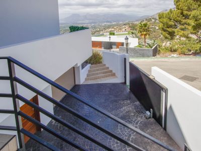 Modern exterior staircase with black railings descends to a textured dark gray driveway, featuring an an automatic gate, garage entrance, and panoramic mountain views.