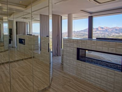Modern interior featuring a transparent gas fireplace with pebble base, set within a textured wall. Mirrored panels reflect the room and the expansive mountain and sea views through floor-to-ceiling windows.