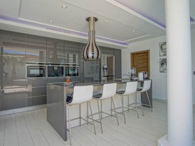 Modern kitchen featuring high-gloss grey cabinetry, integrated ovens, and a central island with seating for four. Recessed and LED strip lighting illuminate the space.