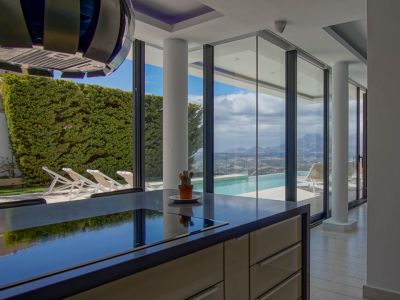Modern kitchen island featuring a dark, reflective countertop with an integrated hob. Floor-to-ceiling glass doors open to an infinity pool and panoramic mountain views.