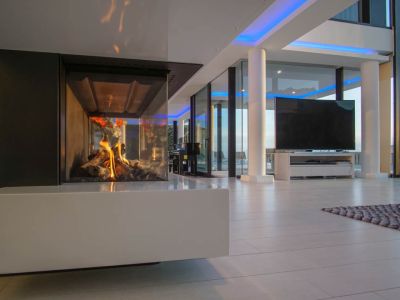 Modern living space featuring a glass-enclosed gas fireplace with a white base. Large windows and blue accent lighting complement the design.