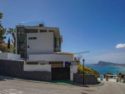Modern white villa exterior with a prominent glass elevator shaft and rooftop terrace railing. Features stone retaining walls, an automatic gate, and panoramic sea views of the coastline.