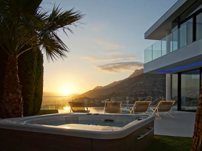 Outdoor jacuzzi with wood-like finish, set on green grass, facing a modern villa with glass balconies. Features sunset mountain and sea views.