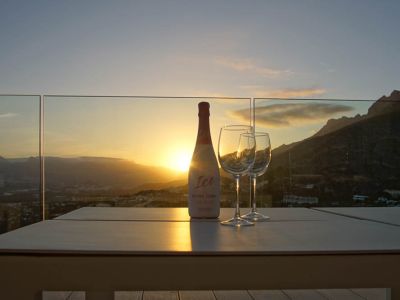 Outdoor table on a terrace with glass balustrade, featuring a rosé cava bottle and two glasses. Sunset over mountains and valley, with warm reflections.
