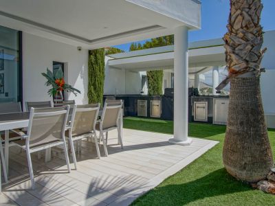 Outdoor terrace with al fresco dining on light plank-style tiling, bordered by artificial grass. Features a covered outdoor kitchen with BBQ and stainless steel appliances, beside a large palm tree.