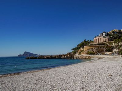 Pebble beach with clear turquoise waters and a rocky coastline. Modern buildings with reflective facades and a rounded structure line the adjacent hillside, all under a bright blue sky.