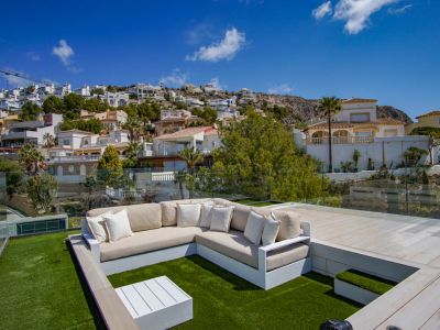 Rooftop lounge area featuring an L-shaped sofa on artificial turf, complemented by a white coffee table and glass railings. Offers clear views of the terraced hillside with residential homes.