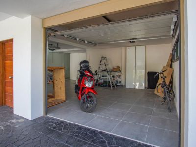 Spacious garage featuring grey tiled flooring, a partially open sectional door, and a distinct wooden entrance door on the left. Inside, a red scooter and bicycle are visible.