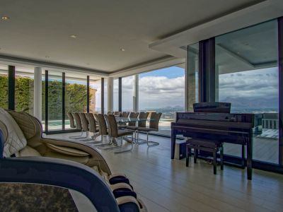 Spacious living and dining area featuring a glass table for ten, a grand piano, and a massage chair. Expansive windows provide panoramic mountain and landscape views.