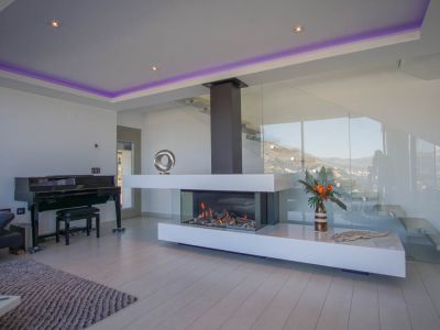 The elegant living area features a modern transparent gas fireplace, a grand piano, and a sleek glass staircase, all set against mountain views.