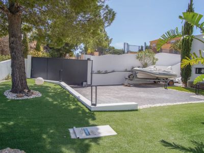 The main entrance features a dark automatic gate and a paved parking area, currently accommodating a boat on a trailer. Artificial grass and a mature pine tree define the foreground.