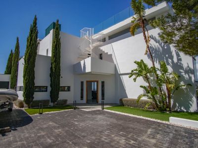 The modern white villa exterior features a paved driveway, tall cypress trees, and a distinctive spiral staircase leading to an upper terrace with glass balustrade.