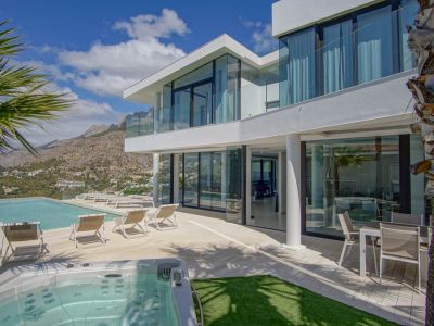 The modern white villa exterior showcases an infinity pool, separate jacuzzi, and a spacious terrace with loungers, all framed by dramatic mountain vistas under a clear sky.