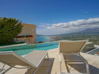 The outdoor terrace features an infinity pool overlooking the coastline and distant mountains. Two sun loungers rest on light decking, framed by a glass railing and a traditional villa facade.
