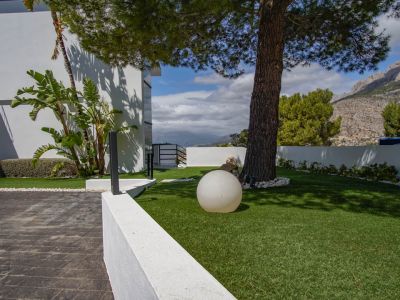 The villa's exterior features a paved entrance leading to an artificial lawn with a mature pine tree, a modern spherical light, and distant mountain views.