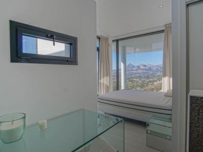This bright bedroom features a bed positioned before expansive floor-to-ceiling windows, offering panoramic mountain and valley views. A glass console and a dark-framed horizontal window are also visible.