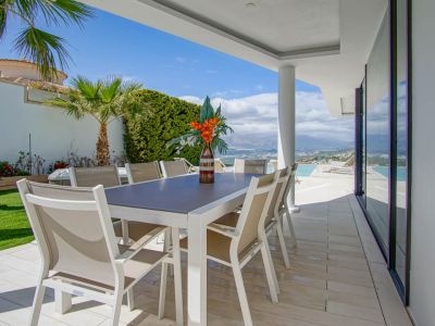 This covered outdoor dining terrace features a modern white table with eight beige chairs, set on light tiles. It offers expansive views of the distant mountains and sea, adjacent to the infinity pool.