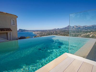 This infinity pool features clear blue water, light decking, and a glass barrier, offering panoramic views of the sea, coastline, and distant mountains under a clear sky.