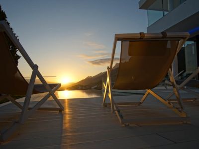 Two modern lounge chairs on light decking overlook an infinity pool, reflecting a golden sunset over distant mountains.