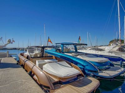 Two modern powerboats, one bronze with a teak deck and another metallic blue, docked in a marina under a clear sky. Spanish flags are visible among the background yachts.
