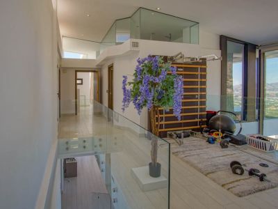 Upper floor gym area featuring modern glass railings, light wood-effect flooring, and a wall-mounted exercise ladder. Panoramic views of the surrounding landscape are visible through expansive floor-to-ceiling windows.