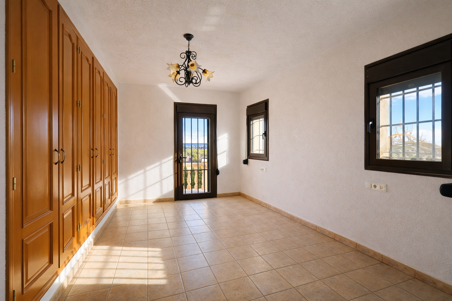 Bright room featuring extensive built-in wooden wardrobes, light tiled flooring, and textured white walls. Includes a balcony door with security bars, offering landscape views.