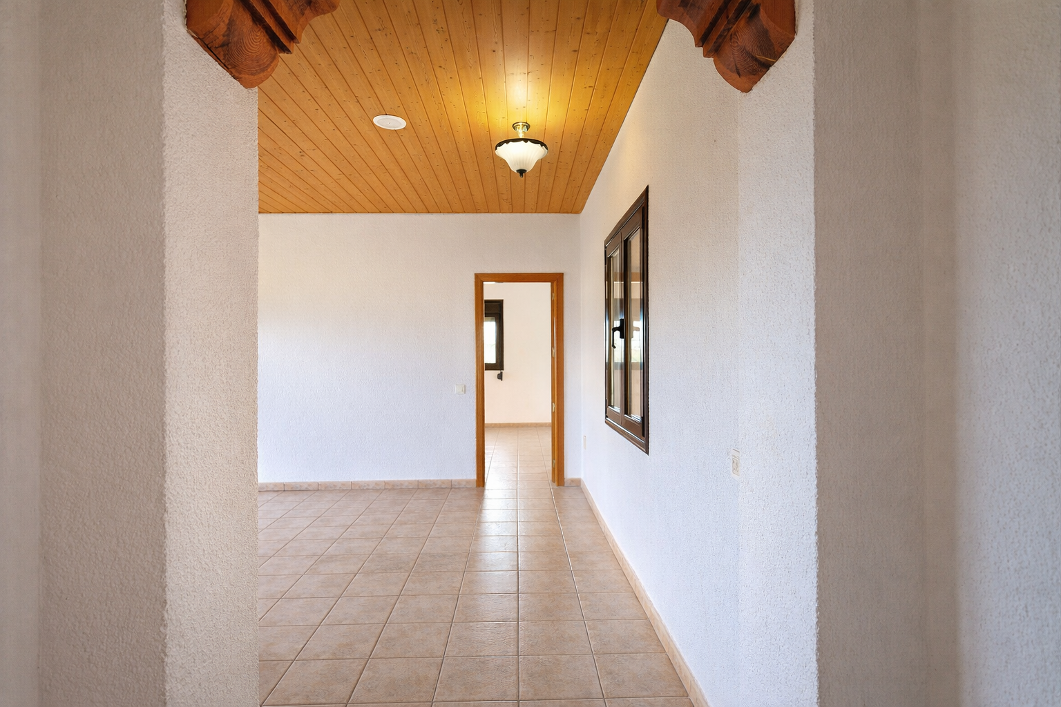 Interior hallway featuring light tiled flooring and textured white walls. Wooden plank ceiling with a central light and carved decorative arch details.