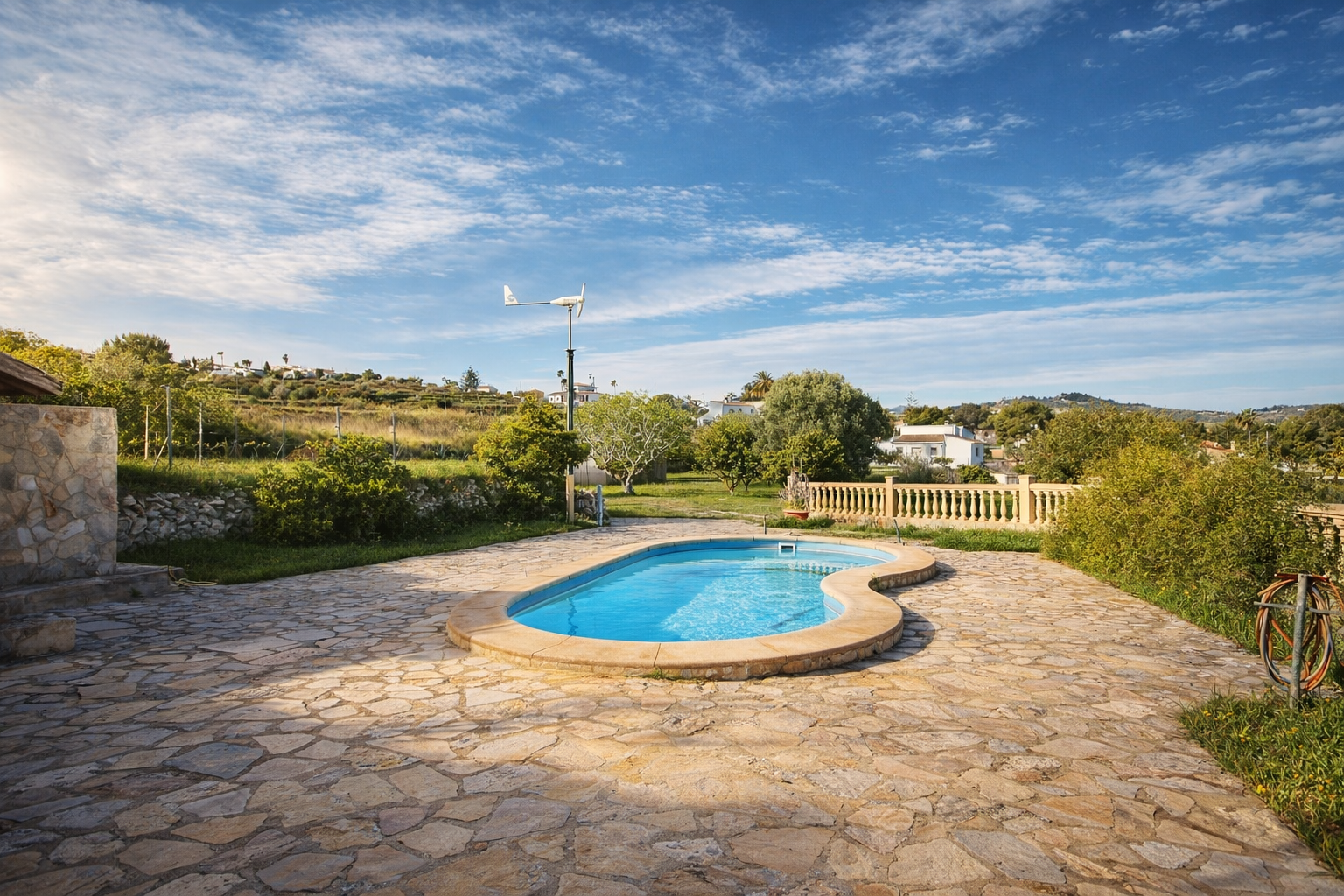 Organic-shaped swimming pool set on an expansive irregular flagstone terrace. Green hills, lush vegetation, and a small wind turbine are visible in the background.