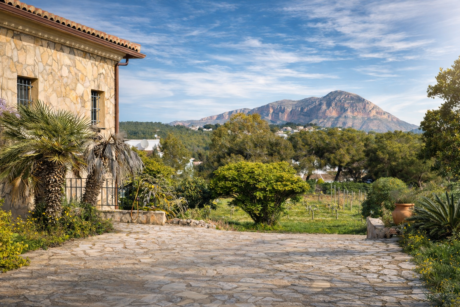 Rustic stone facade of the finca with palm trees, flagstone driveway, and views of Montgó, surrounded by vineyards and Mediterranean vegetation.