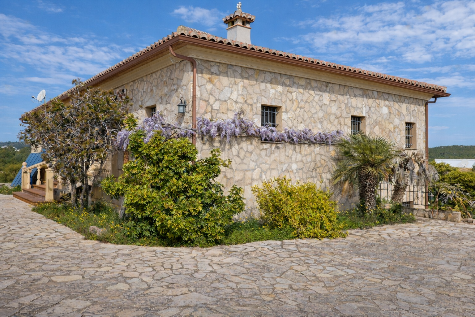 Rustic stone facade with a tiled roof, adorned by purple wisteria below the grilled windows. The entrance path is paved with irregular stones.