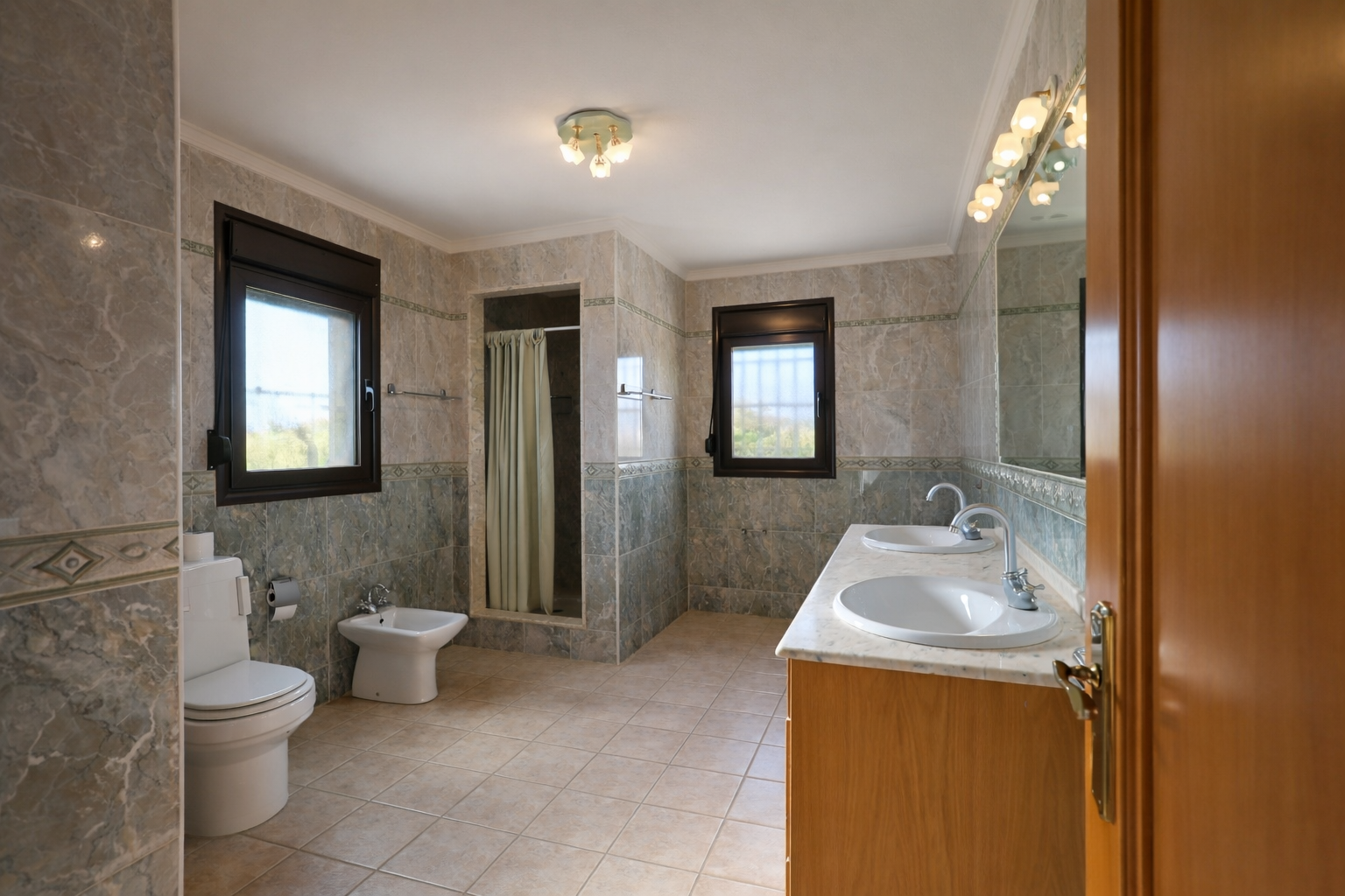Spacious bathroom featuring a double vanity with marble countertop and wooden cabinet. Includes a walk-in shower, toilet, bidet, and two windows providing natural light.