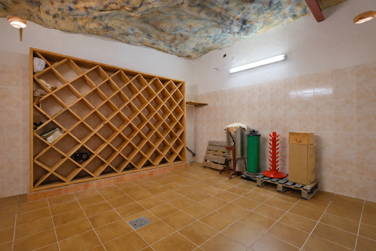 Spacious cellar featuring a large wooden grid wine rack, tiled walls, and ceramic floor. A textured cave-like ceiling and functional lighting are visible.