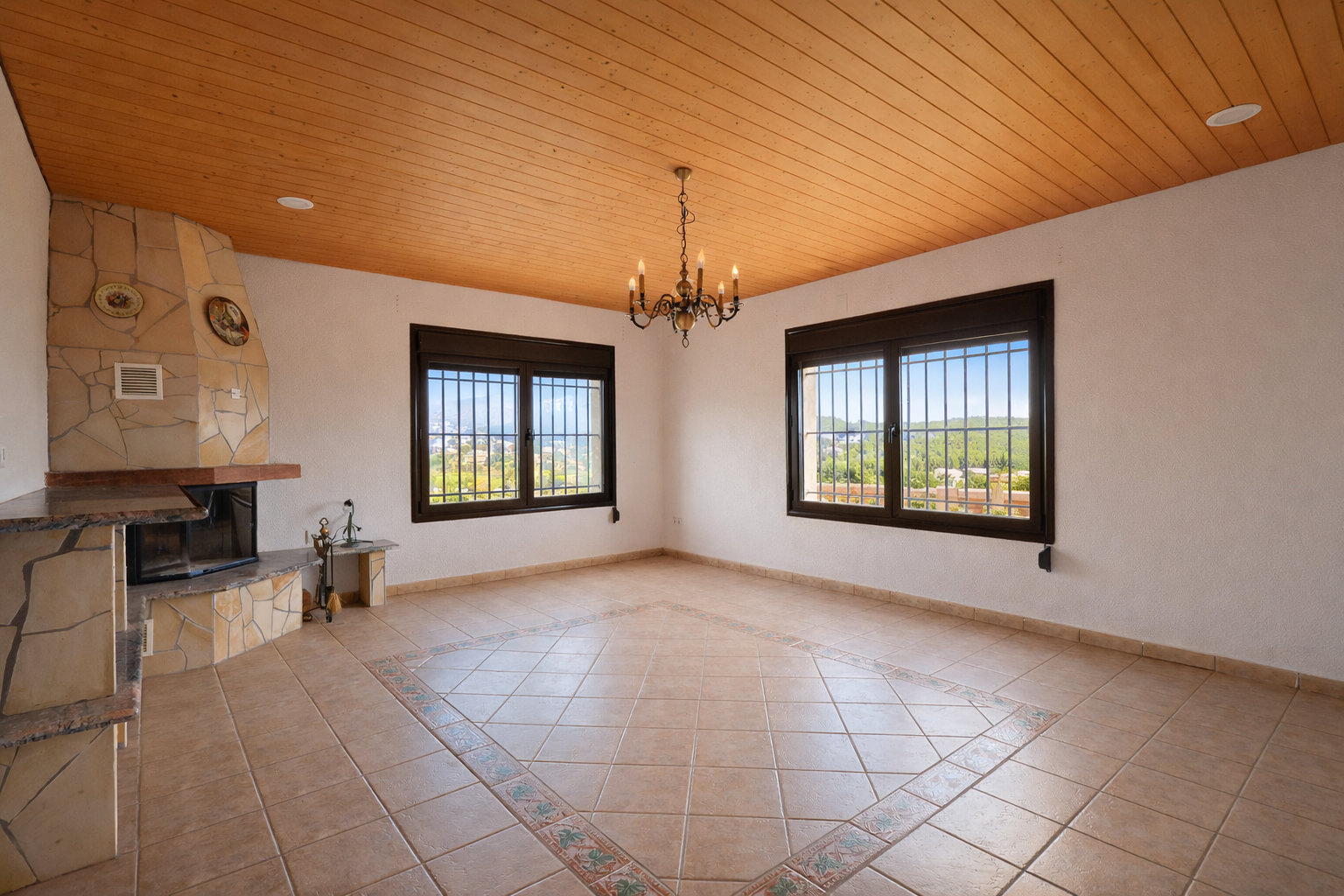 Spacious room featuring a stone-clad fireplace, a wooden plank ceiling, and a tiled floor with a decorative central pattern. Two barred windows offer valley views.