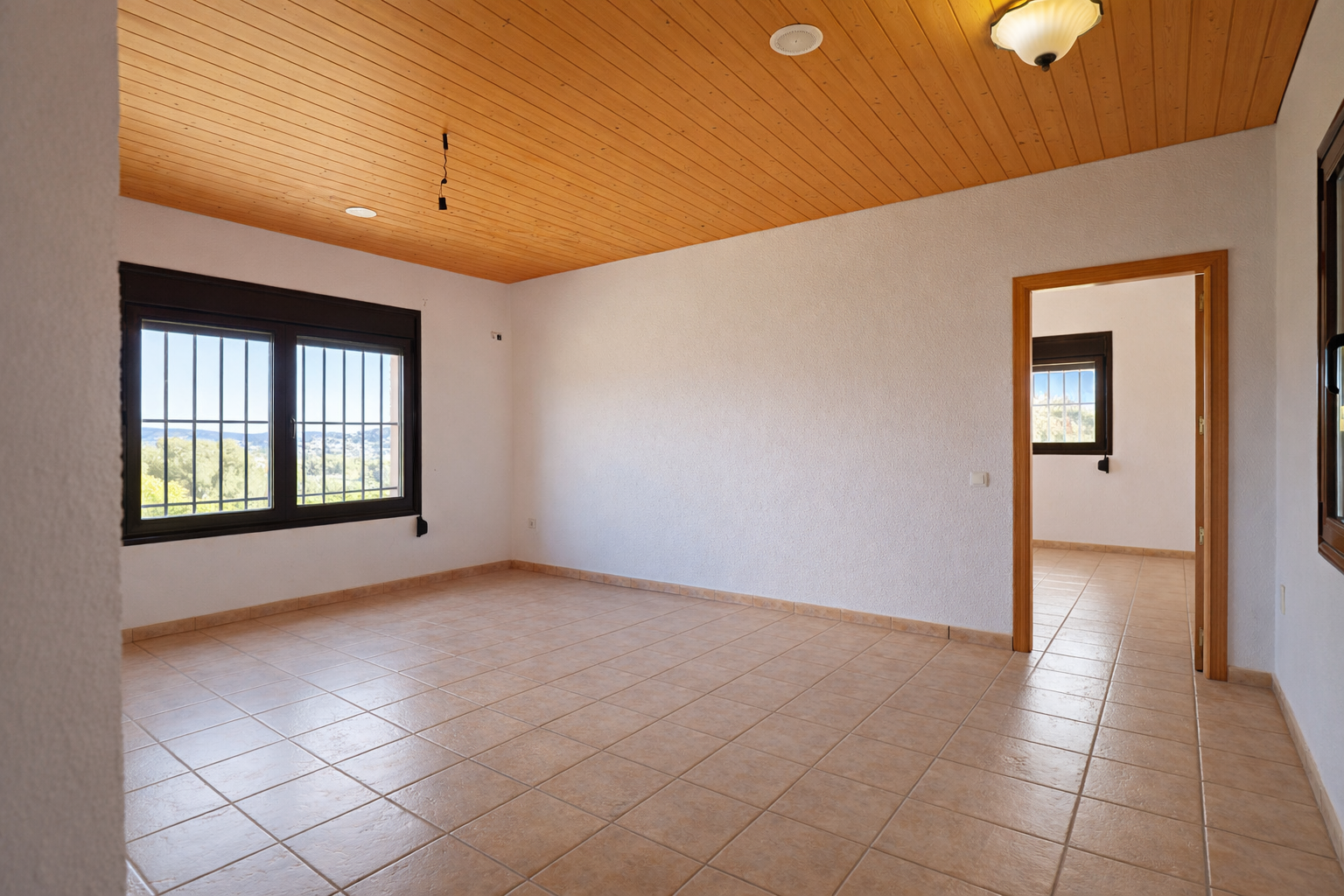 Spacious room featuring light tiled flooring, white textured walls, and a warm wooden slatted ceiling. Windows with security bars provide natural light and valley views.