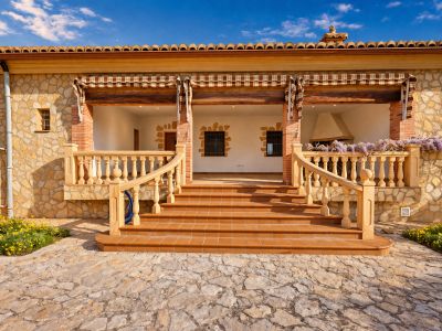 Main entrance featuring a spacious naya with terracotta flooring, natural stone facade, and a classic balustrade staircase. An outdoor fireplace, wisteria, and a solar panel are visible.