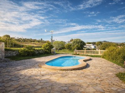 Organic-shaped swimming pool set on an expansive irregular flagstone terrace. Green hills, lush vegetation, and a small wind turbine are visible in the background.