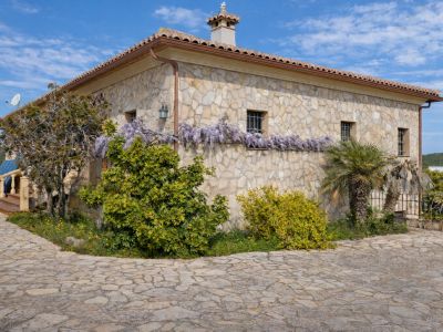 Rustic stone facade with a tiled roof, adorned by purple wisteria below the grilled windows. The entrance path is paved with irregular stones.