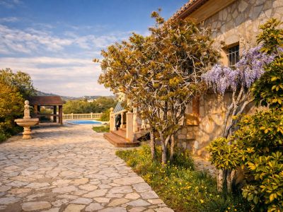 Stone facade of the finca with blooming wisteria. A flagstone path leads to the pool area and a wooden pergola, featuring a stone fountain in the garden.