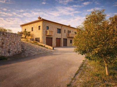 The stone-clad exterior of the finca features three integrated garages and elegant balconies, accessed via a paved driveway and a curved stone wall.