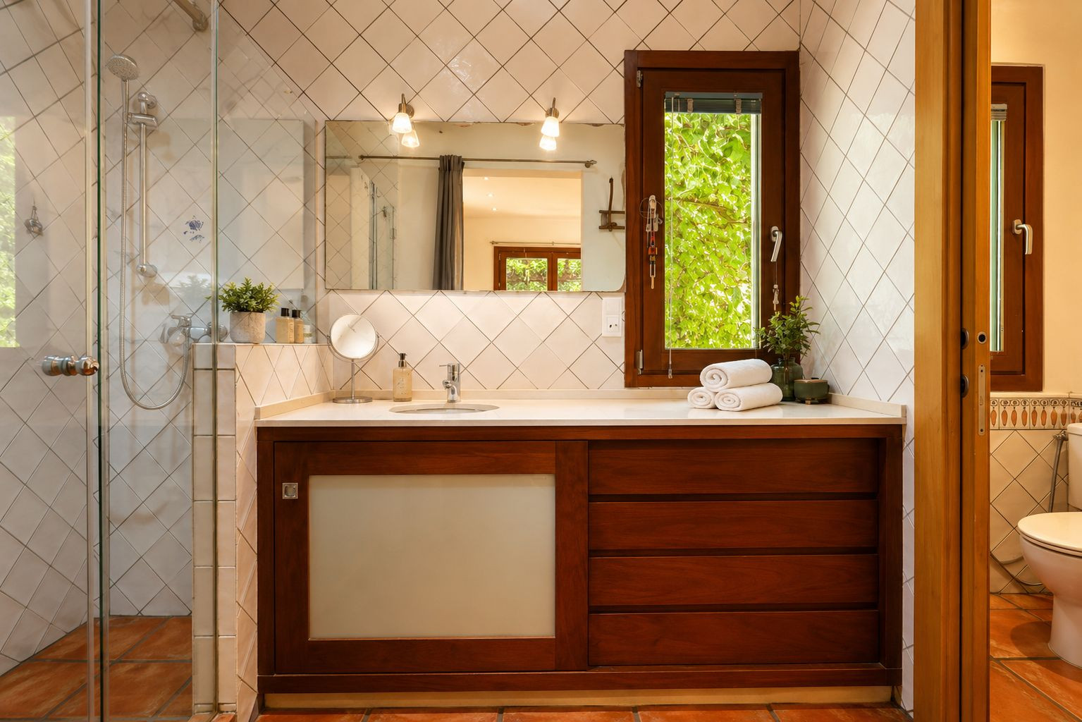 Bathroom featuring a glass shower, dark wood vanity with white countertop and integrated sink. A window offers green views, complemented by diamond-patterned white tiles and terracotta floors.