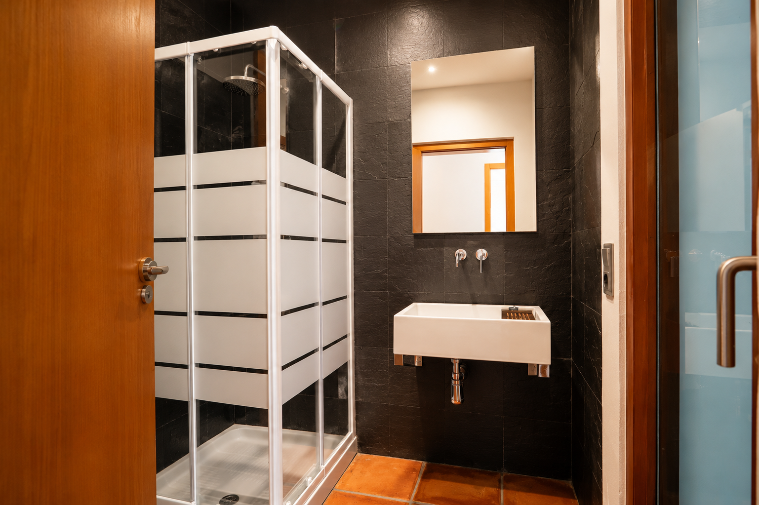 Bathroom featuring an enclosed shower with frosted stripes, a wall-mounted rectangular basin, and dark textured wall tiles. Terracotta floor tiles complete the space.