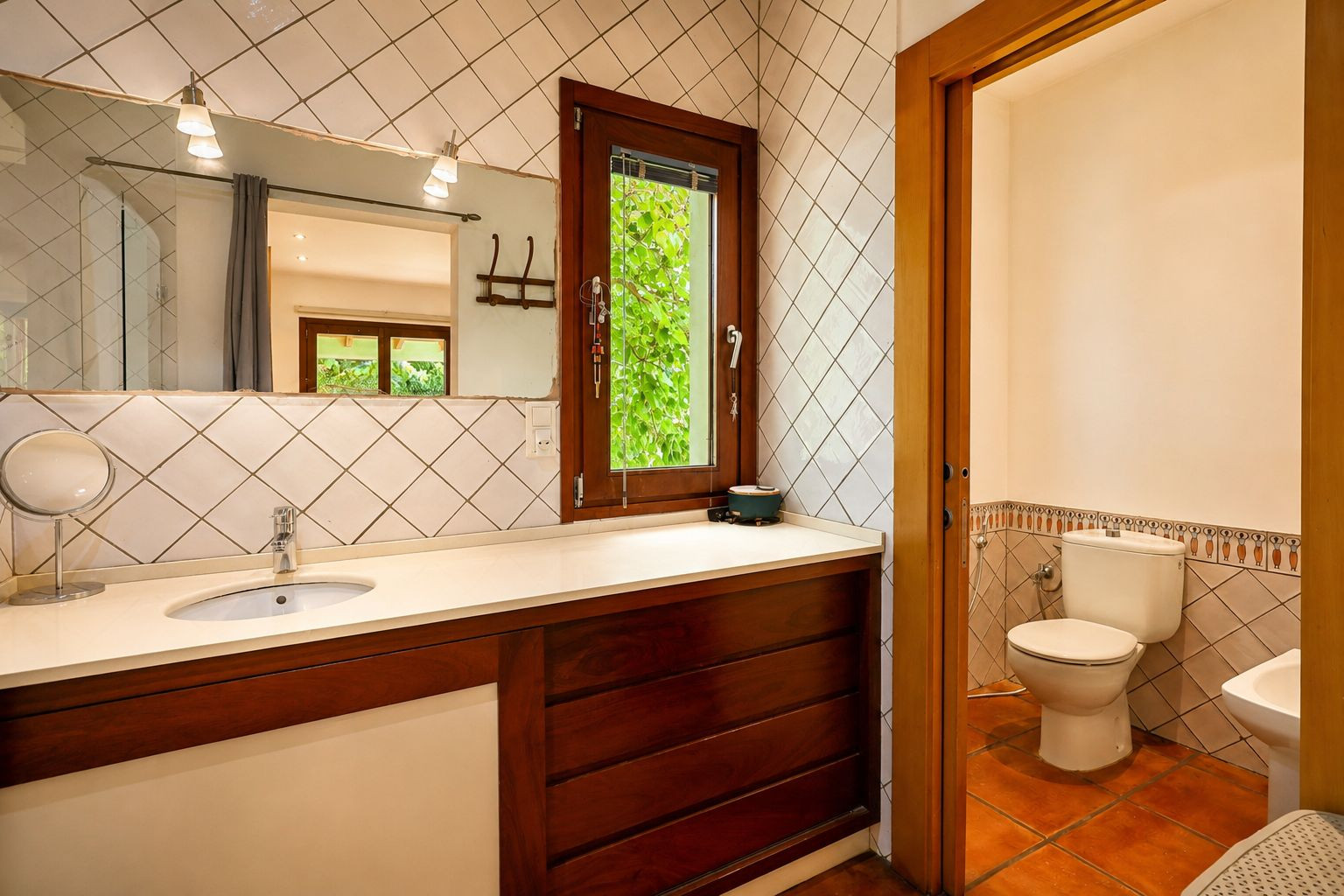 Bathroom vanity featuring a single sink set in a light countertop, dark wooden cabinet, and a large mirror with sconce lighting. A tall window frames lush green foliage outside.