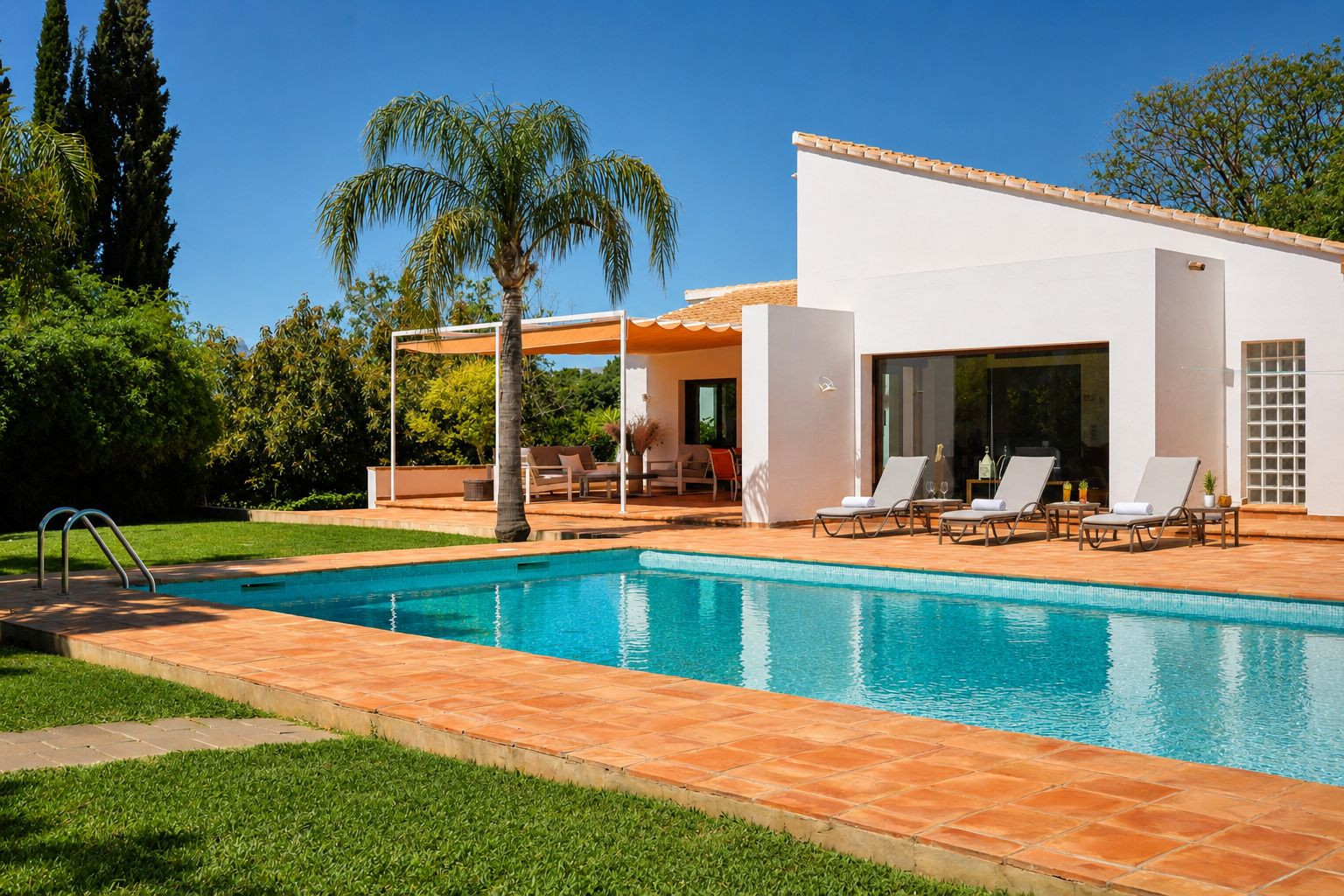 Blue-tiled swimming pool with terracotta surround, set amidst green lawn. Sun loungers line the white main house façade, adjacent to a shaded pergola with outdoor lounge furniture.
