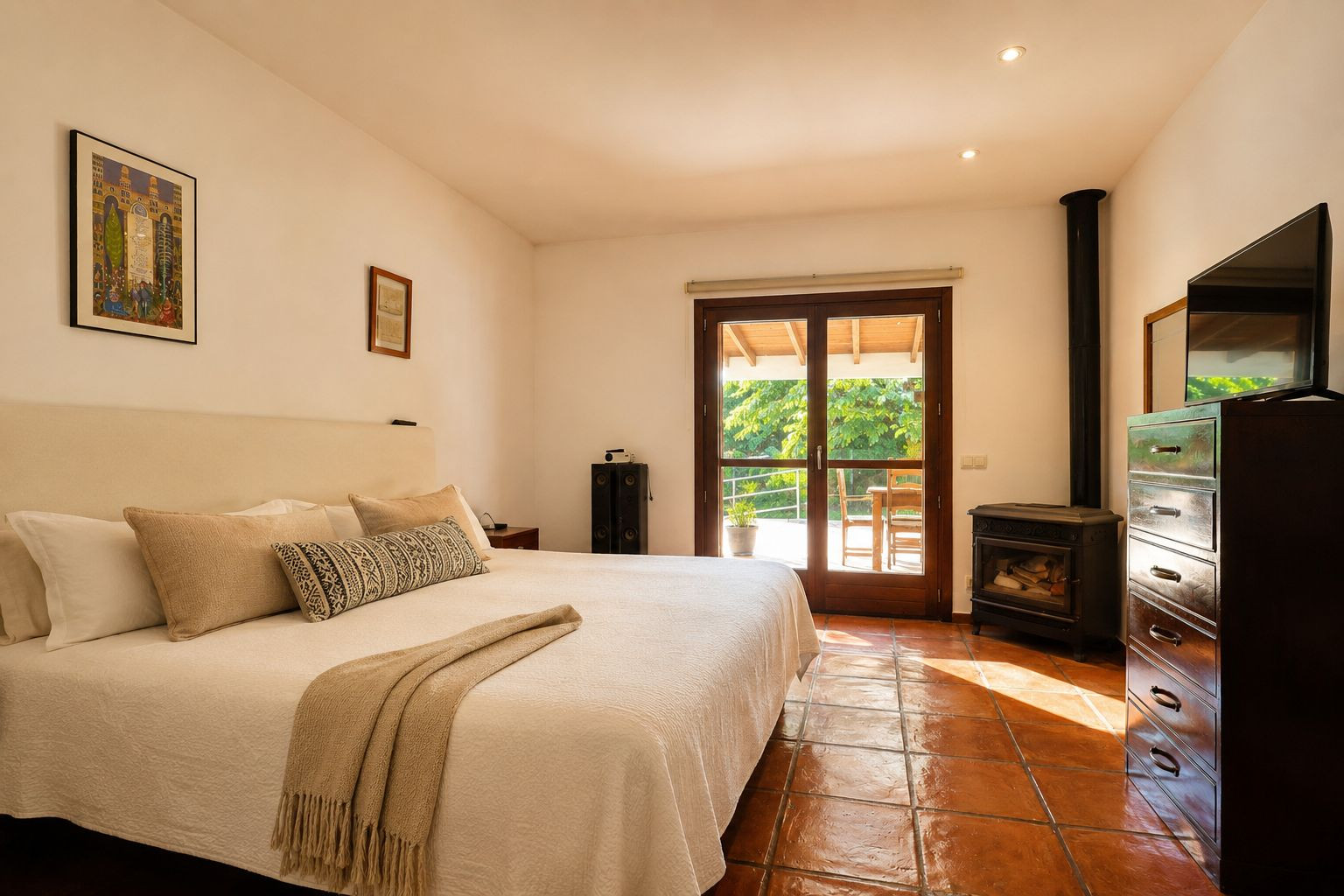 Bright bedroom featuring a large bed, traditional terracotta floor tiles, and a cozy wood-burning stove. Double glass doors open to a private outdoor terrace amidst lush greenery.